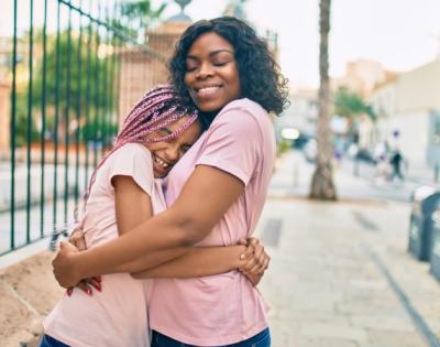 Mom hugs her daughter while standing on a sidewalk. Both are wearing pink shirts.