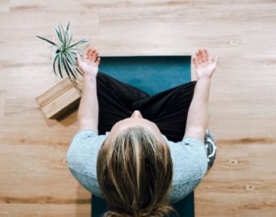 person sitting on blue yoga mat in cross-legged position on wooden floor with plant and yoga blocks next to them