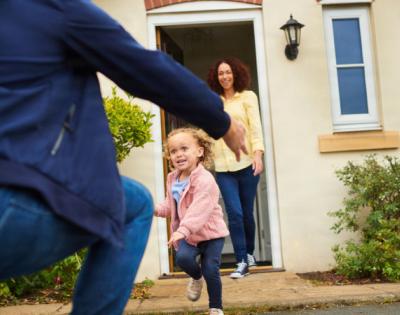 Child running to father with mother standing and smiling in the house doorway behind the child.