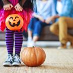 Child in costume holding a pumpkin basket with parents sitting on couch in background